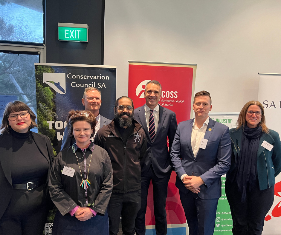 Event faciliator, Dr Gemma Beale, AICSA Co-Chair Emma Webb, SACOSS CEO Ross Womersley, Kuma Kaaru's Cliffy Wilson, The Hon Peter Malinauskas MP, Premier of South Australia, SA Unions Secretary Dale Beasley and Conservation Council SA CEO Kirsty Bevan pictured smiling, standing in front of organisation banners.